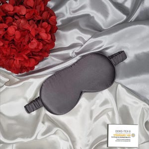 May include: A gray silk sleep mask with an elastic band. The mask is on a white satin surface. The text "OEKO-TEX&reg; CONFIDENCE IN TEXTILES STANDARD 100 13.HTR.01133  Hohenstein HTTI Tested for harmful substances www.oeko-tex.com/standard100" is printed on a white label.