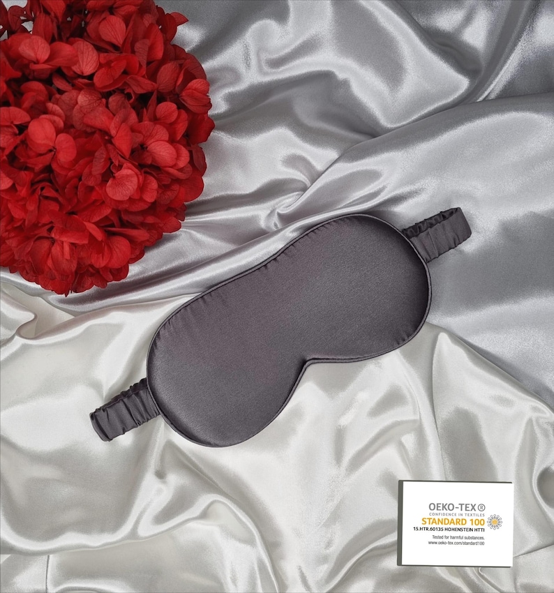 May include: A gray silk sleep mask with an elastic band. The mask is on a white satin surface. The text "OEKO-TEX&reg; CONFIDENCE IN TEXTILES STANDARD 100 13.HTR.01133  Hohenstein HTTI Tested for harmful substances www.oeko-tex.com/standard100" is printed on a white label.