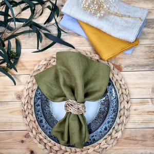 May include: A set of linen napkins in olive green, mustard yellow, light blue, and light gray, with one olive green napkin folded and secured by a woven rope napkin ring on a speckled ceramic plate and a braided round placemat.