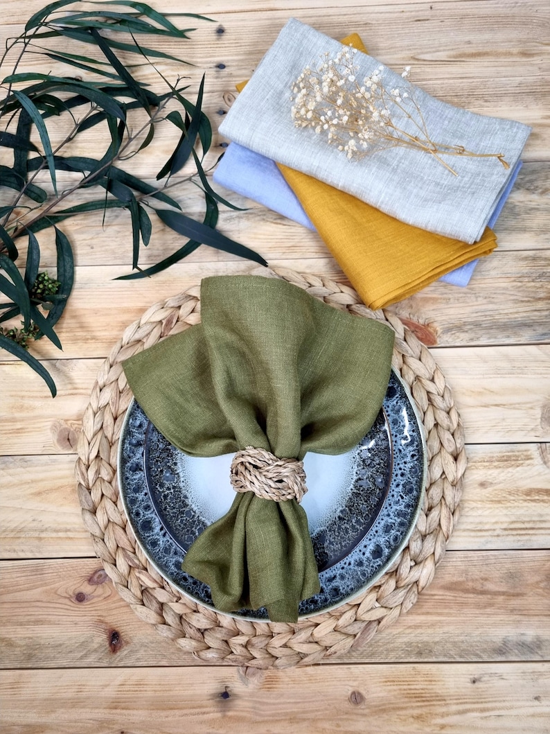 May include: A set of linen napkins in olive green, mustard yellow, light blue, and light gray, with one olive green napkin folded and secured by a woven rope napkin ring on a speckled ceramic plate and a braided round placemat.
