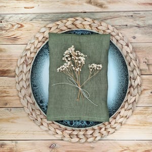 May include: A rectangular olive green linen napkin folded over a round blue and white speckled ceramic plate, which rests on a circular woven straw placemat. A small, delicate sprig of dried white baby's breath flowers is tied with white string.