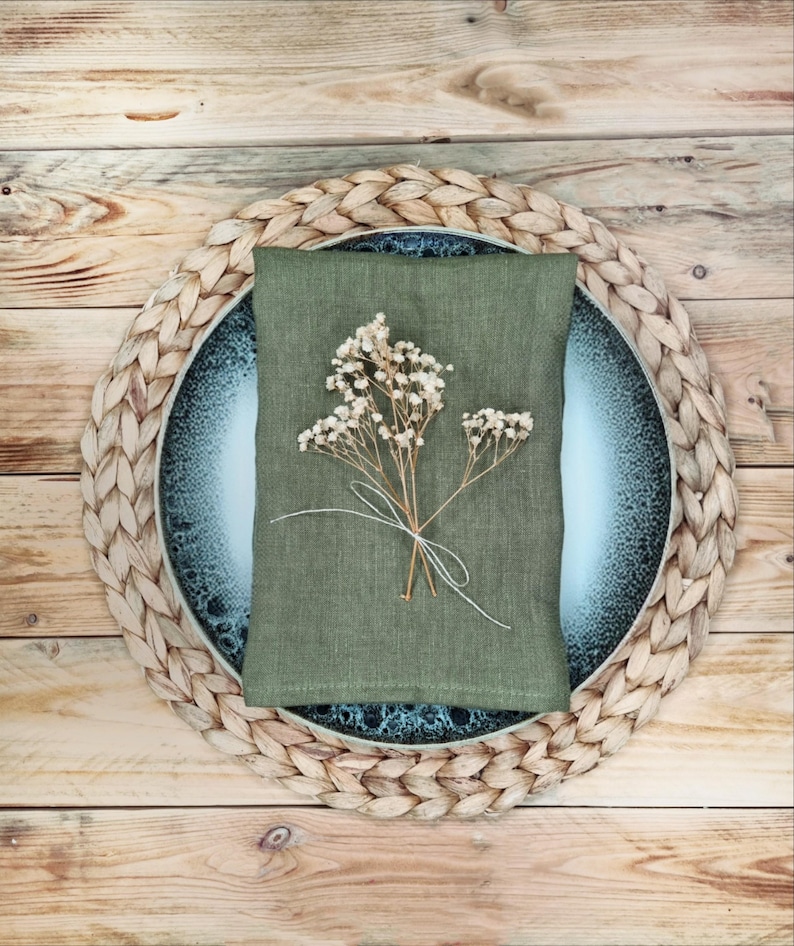 May include: A rectangular olive green linen napkin folded over a round blue and white speckled ceramic plate, which rests on a circular woven straw placemat. A small, delicate sprig of dried white baby's breath flowers is tied with white string.