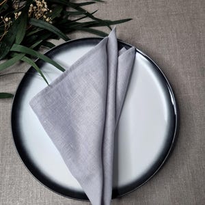 May include: A light gray linen napkin folded into a triangle shape, placed on a round ceramic plate with a white center and a dark charcoal gray rim. The napkin features a visible woven texture and clean stitched edges, creating a simple display.