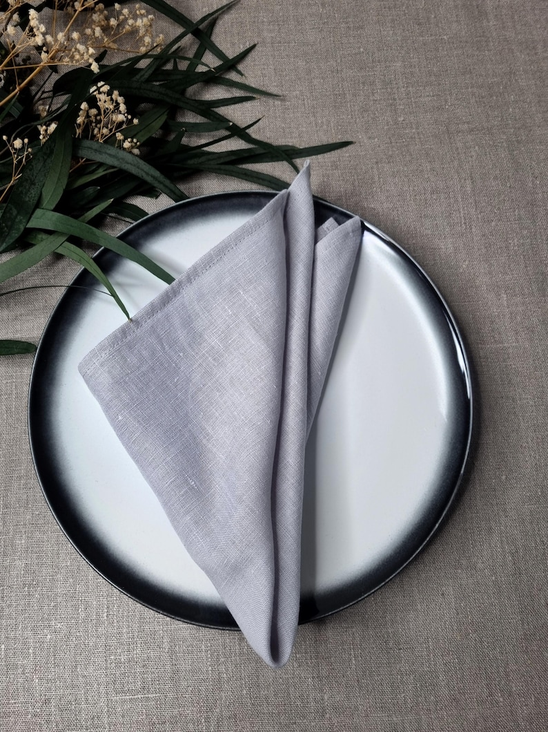 May include: A light gray linen napkin folded into a triangle shape, placed on a round ceramic plate with a white center and a dark charcoal gray rim. The napkin features a visible woven texture and clean stitched edges, creating a simple display.