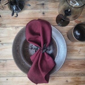 May include: A deep burgundy linen napkin folded and placed inside a round, speckled grey ceramic plate. The napkin is secured with a small, dark sprig of eucalyptus, creating a textured and layered appearance on the dinnerware piece.