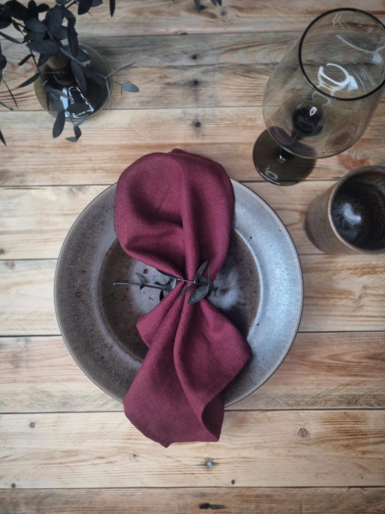 May include: A deep burgundy linen napkin folded and placed inside a round, speckled grey ceramic plate. The napkin is secured with a small, dark sprig of eucalyptus, creating a textured and layered appearance on the dinnerware piece.
