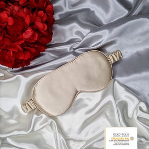 May include: A beige silk sleep mask with an elastic band. The mask is on a white satin background. The text on the label reads "OEKO-TEX&reg; CONFIDENCE IN TEXTILES STANDARD 100 13.HTR.40133 HOHENSTEIN HTTI Tested for harmful substances www.oeko-tex.com/standard100"