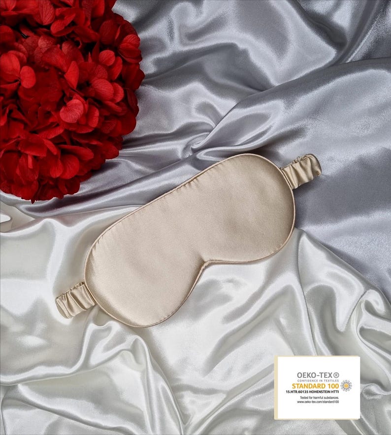 May include: A beige silk sleep mask with an elastic band. The mask is on a white satin background. The text on the label reads "OEKO-TEX&reg; CONFIDENCE IN TEXTILES STANDARD 100 13.HTR.40133 HOHENSTEIN HTTI Tested for harmful substances www.oeko-tex.com/standard100"