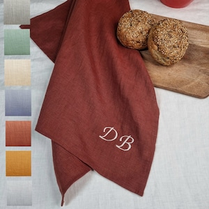 May include: A red linen kitchen towel with the initials "DB" embroidered in white. The towel is folded and placed on a white linen tablecloth. Two round rolls of bread are on a wooden chopping board.