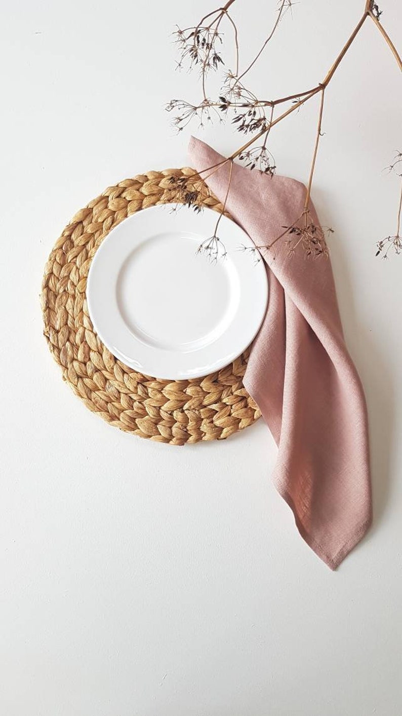 May include: A round, woven natural fiber placemat topped with a plain white ceramic plate and a folded dusty pink linen napkin. A delicate, dried brown plant stem with small seed heads extends over the top of the plate and napkin arrangement.