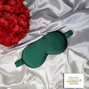 May include: A dark green silk sleep mask on a white satin background. The mask has a black elastic band and is labeled with the Oeko-Tex Standard 100 logo.