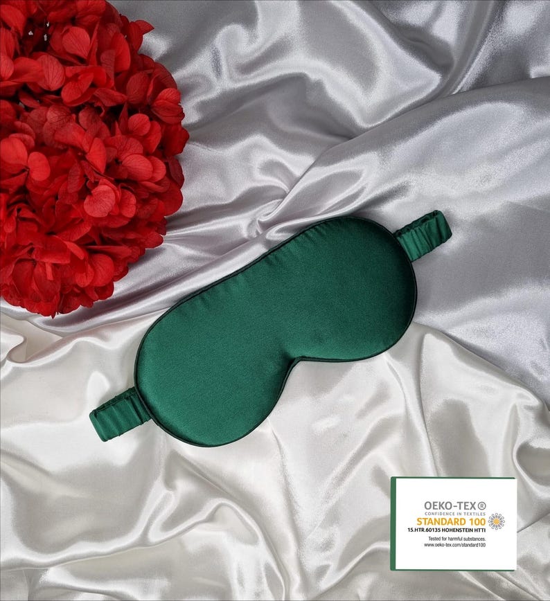 May include: A dark green silk sleep mask on a white satin background. The mask has a black elastic band and is labeled with the Oeko-Tex Standard 100 logo.