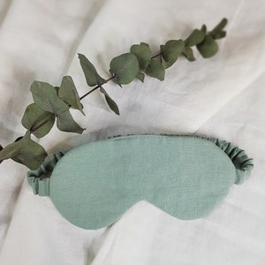 Puede incluir: Una m&aacute;scara para dormir de color verde claro hecha de lino con un borde rizado. La m&aacute;scara est&aacute; sobre un fondo de tela blanca.