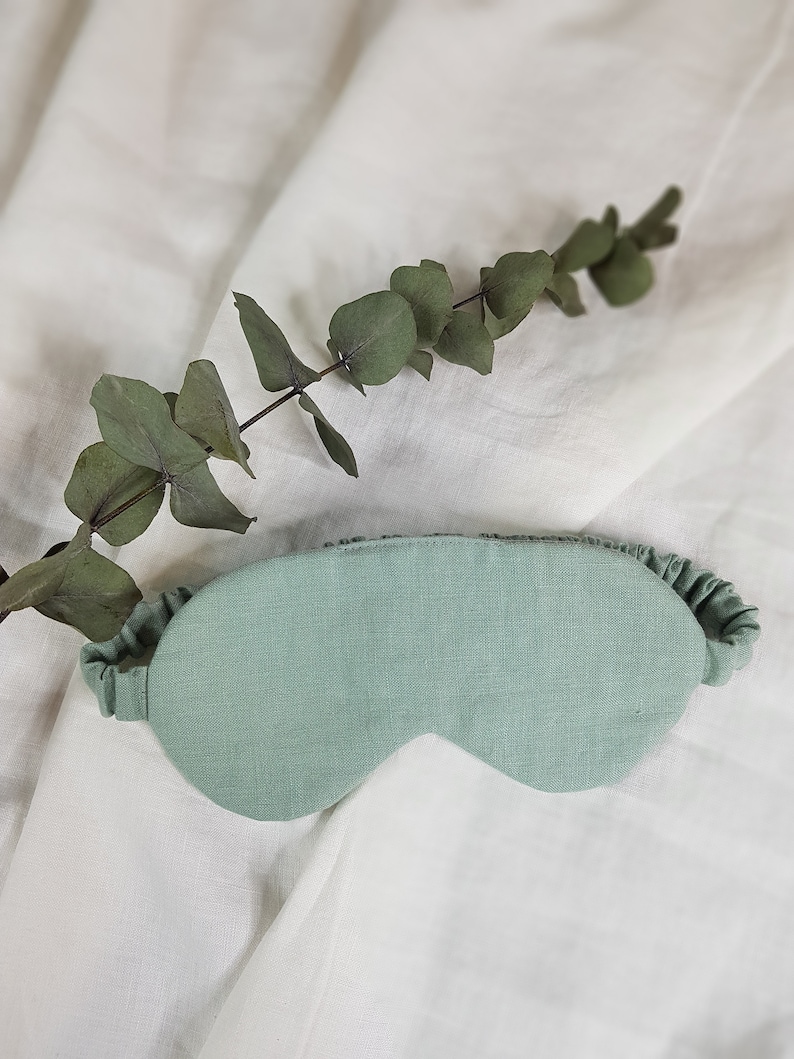 Puede incluir: Una m&aacute;scara para dormir de color verde claro hecha de lino con un borde rizado. La m&aacute;scara est&aacute; sobre un fondo de tela blanca.