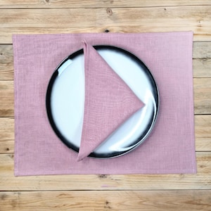 May include: A rectangular dusty rose linen placemat featuring a matching triangular folded napkin placed on top of a round white plate with a wide black rim. The plate and napkin are centered on the fabric placemat, showcasing a textured weave.