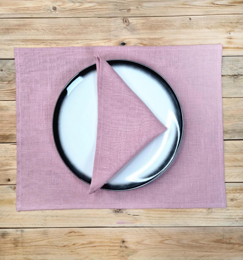 May include: A rectangular dusty rose linen placemat featuring a matching triangular folded napkin placed on top of a round white plate with a wide black rim. The plate and napkin are centered on the fabric placemat, showcasing a textured weave.