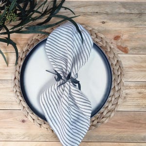 May include: A white and gray striped linen napkin folded and secured with a dark green leaf-shaped napkin ring, placed on a round white plate with a dark gray rim, which sits atop a circular woven braided placemat.