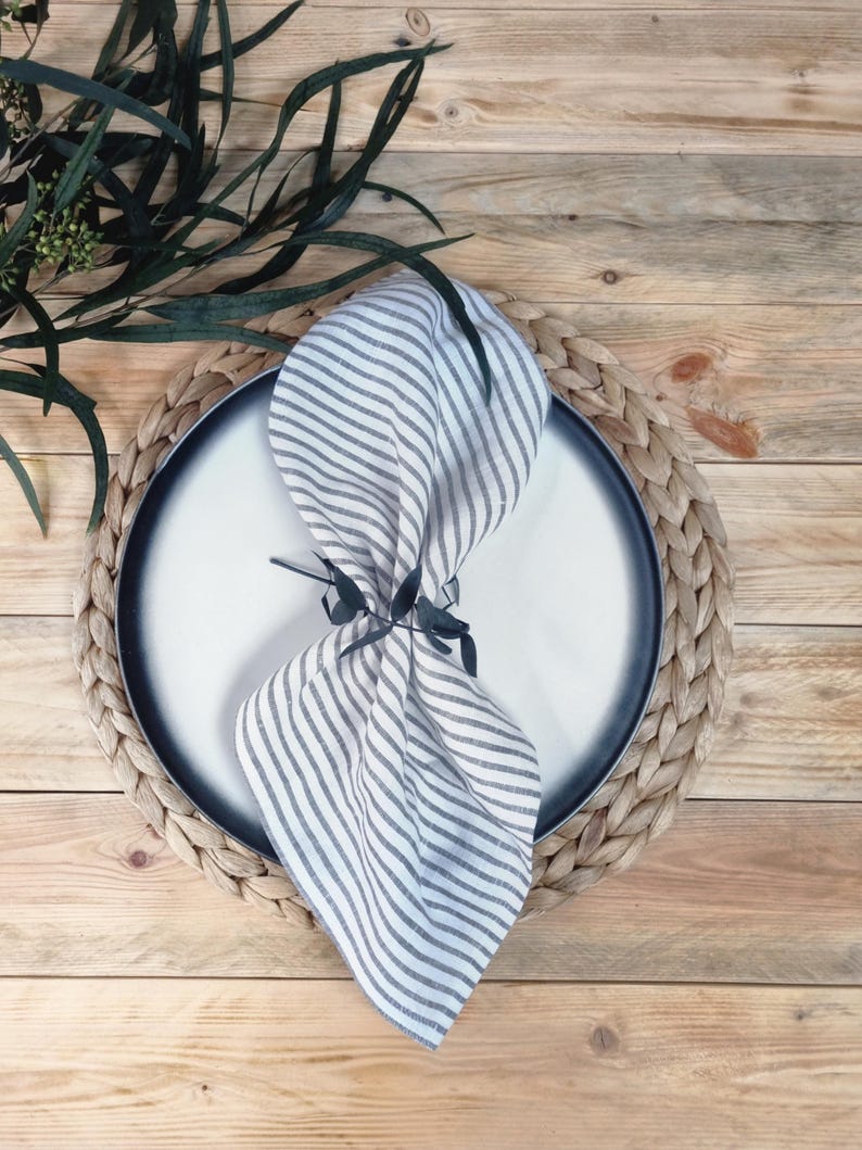 May include: A white and gray striped linen napkin folded and secured with a dark green leaf-shaped napkin ring, placed on a round white plate with a dark gray rim, which sits atop a circular woven braided placemat.