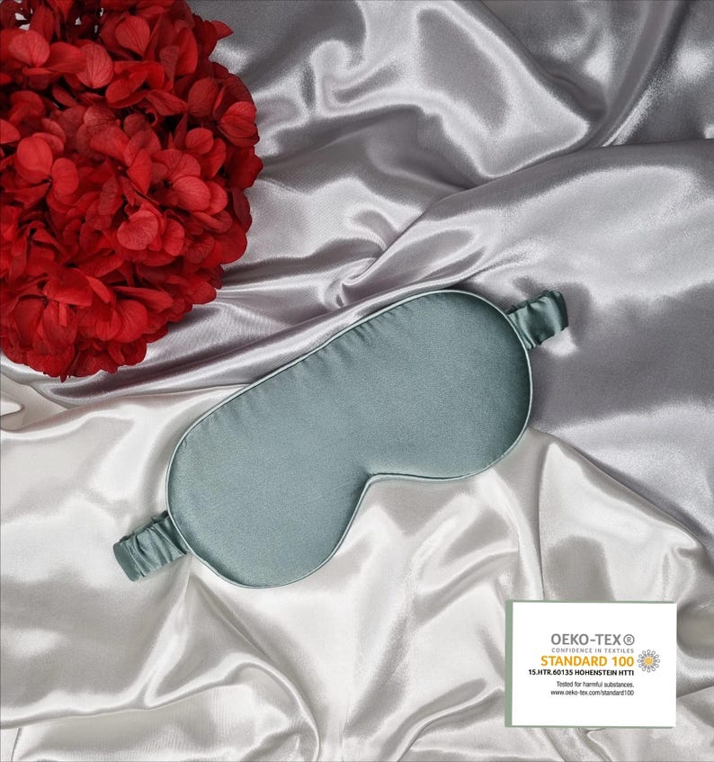 May include: A light blue satin sleep mask with an elastic band. The mask has the OEKO-TEX&reg; STANDARD 100 logo and text: "CONFIDENCE IN TEXTILES  STANDARD 100  13.JATR.90133 HOHENSTEIN HTTI  Tested for harmful substances  www.oeko-tex.com/standard100"