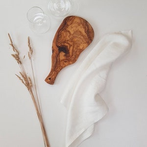 May include: An olive wood serving board with a natural grain pattern, a white linen napkin, two clear glass tumblers, and three dried wheat stalks. The board features a handle with a small hole, and the items are arranged on a plain surface.