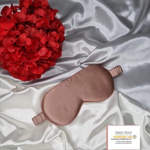 May include: A rose gold satin sleep mask on a white satin background. The sleep mask has a thin elastic band. The background features a red flower. The sleep mask is certified Oeko-Tex Standard 100, which means it is tested for harmful substances.