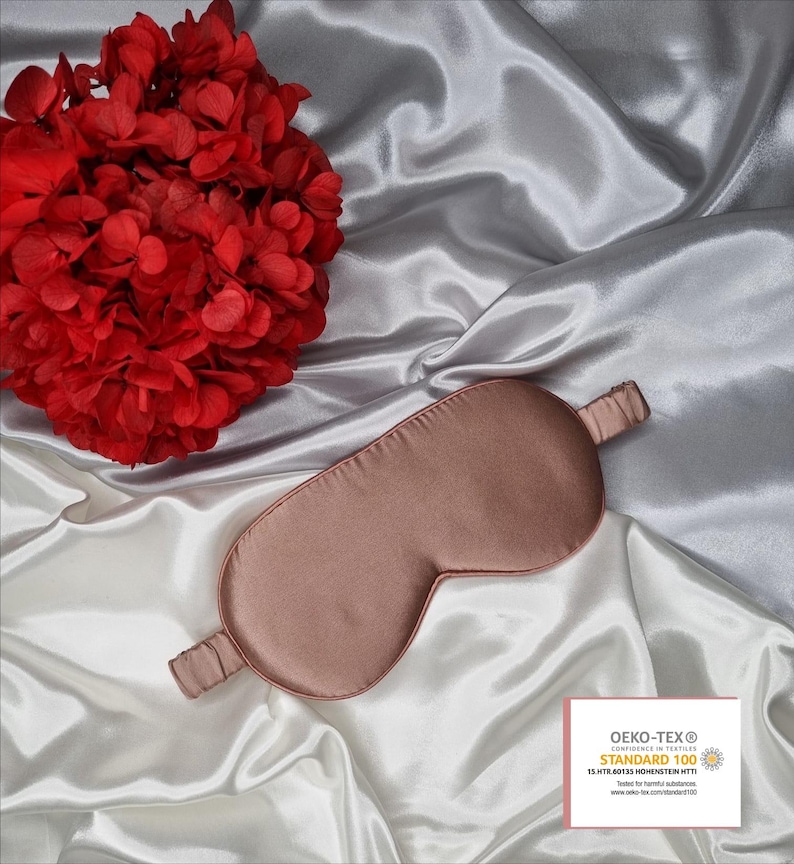 May include: A rose gold satin sleep mask on a white satin background. The sleep mask has a thin elastic band. The background features a red flower. The sleep mask is certified Oeko-Tex Standard 100, which means it is tested for harmful substances.