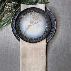 May include: Two round ceramic plates with a reactive glaze featuring a white center and a dark, speckled blue-black rim. A beige linen napkin is folded vertically beneath the plates, and a small sprig of dried white flowers rests on top.