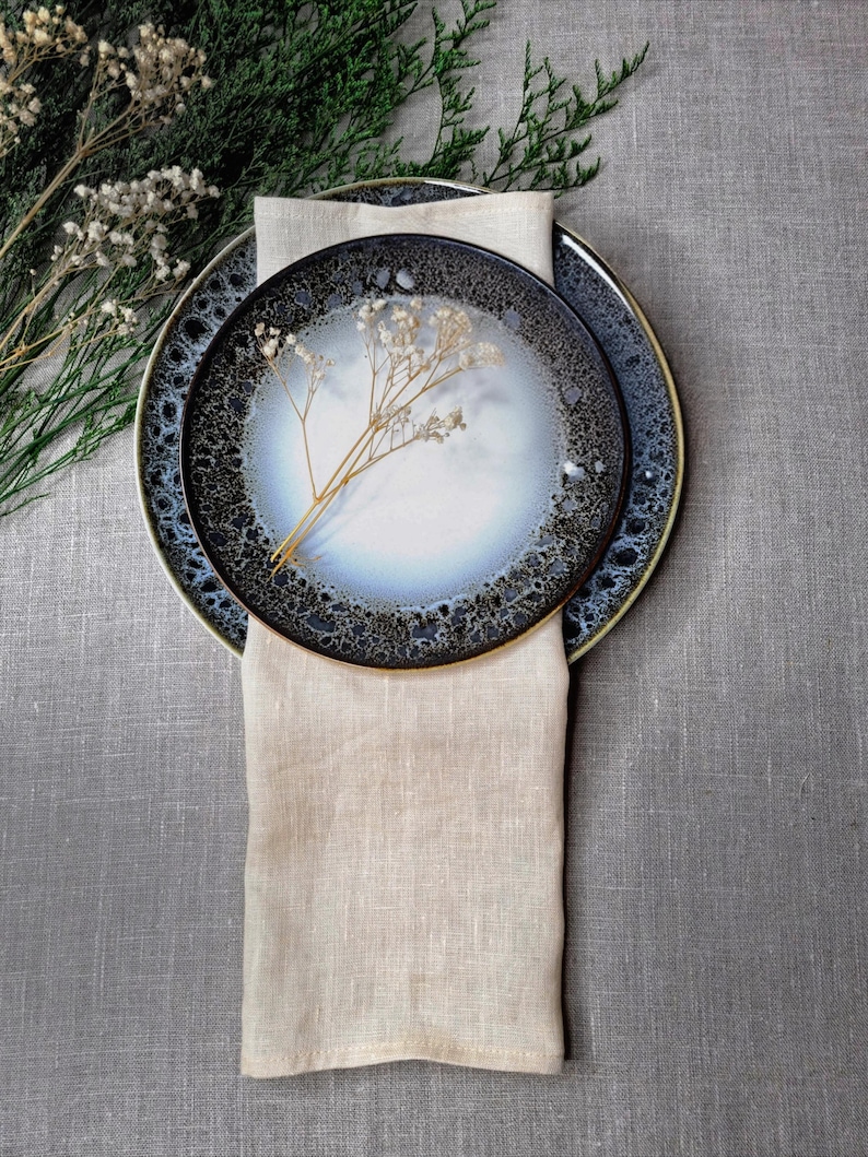 May include: Two round ceramic plates with a reactive glaze featuring a white center and a dark, speckled blue-black rim. A beige linen napkin is folded vertically beneath the plates, and a small sprig of dried white flowers rests on top.
