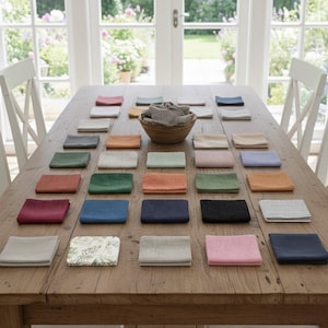 May include: A collection of square fabric napkins in various solid colors and patterns, including shades of blue, green, pink, orange, and grey, arranged on a wooden table with a wooden bowl containing folded napkins in the center.