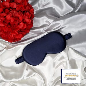 May include: A dark blue silk sleep mask on a white satin background. The mask has a band that goes around the head. The mask has the Oeko-Tex Standard 100 logo on it.