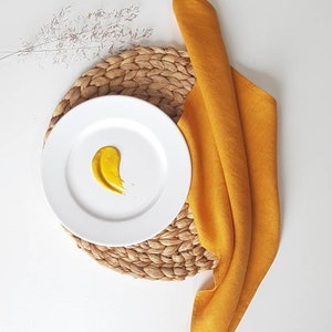 May include: A white ceramic plate featuring a single smear of mustard yellow sauce, placed atop a round woven natural fiber placemat. A mustard yellow fabric napkin is folded and draped alongside the plate, accompanied by a dried grass sprig.