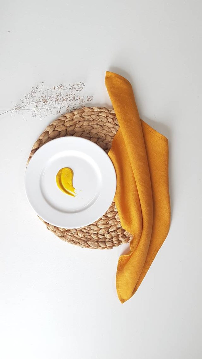 May include: A white ceramic plate featuring a single smear of mustard yellow sauce, placed atop a round woven natural fiber placemat. A mustard yellow fabric napkin is folded and draped alongside the plate, accompanied by a dried grass sprig.