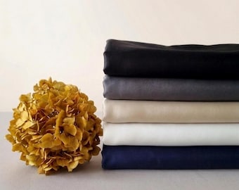 May include: A stack of five folded fabric pieces in black, dark gray, beige, white, and navy blue, positioned next to a dried golden-yellow hydrangea flower head. The textiles appear smooth and opaque, arranged in a neat, horizontal pile.