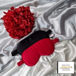 May include: Two silk sleep masks, one black and one red, on a white satin background. The masks are made of soft, comfortable silk and are designed to block out light for a restful sleep. The image also includes a small label with the text "OEKO-TEX&reg; CONFIDENCE IN TEXTILES STANDARD 100 15.HTR.40135 HOHENSTEIN HTTI Tested for harmful substances www.oeko-tex.com/standard100"