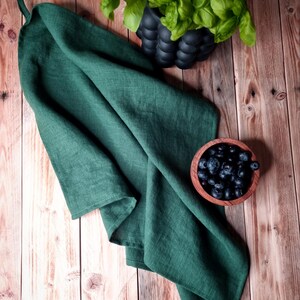 May include: A dark green linen kitchen towel folded on a wooden surface with a bowl of blueberries and a potted plant with green leaves.
