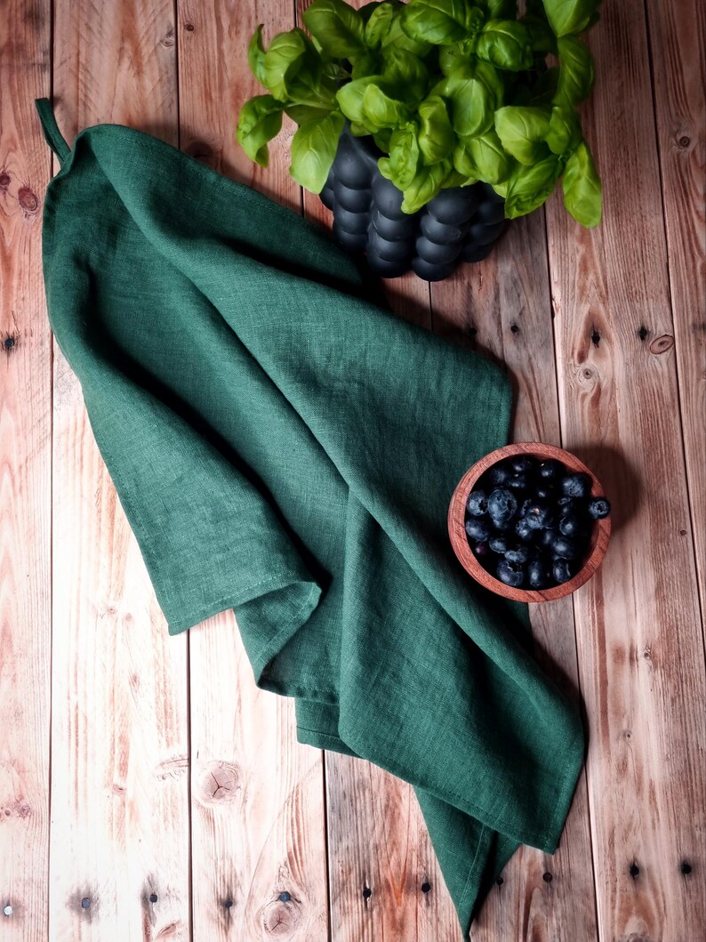 May include: A dark green linen kitchen towel folded on a wooden surface with a bowl of blueberries and a potted plant with green leaves.