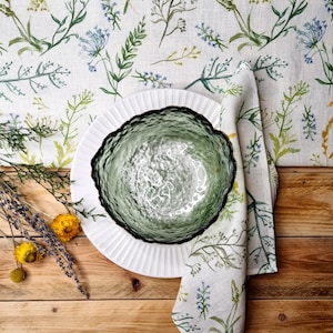 May include: A green textured glass bowl sits on a white fluted plate atop a linen fabric featuring a botanical print of green and blue wildflowers. A matching linen napkin is folded beside the bowl, accompanied by sprigs of dried lavender and yellow flowers.
