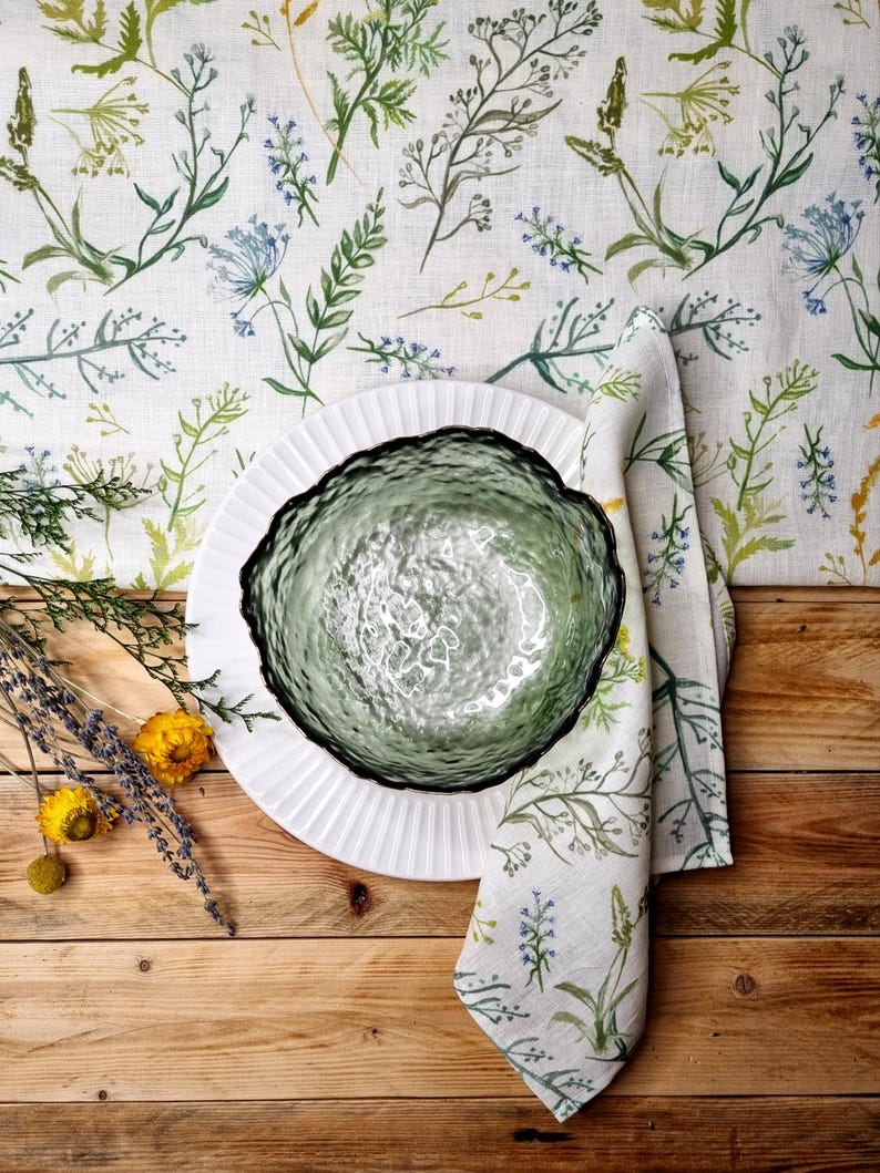 May include: A green textured glass bowl sits on a white fluted plate atop a linen fabric featuring a botanical print of green and blue wildflowers. A matching linen napkin is folded beside the bowl, accompanied by sprigs of dried lavender and yellow flowers.