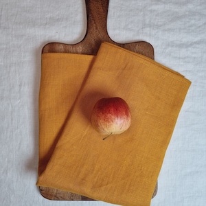 May include: Two mustard yellow linen tea towels folded on a wooden cutting board with a red apple on top. The cutting board has a wooden handle.