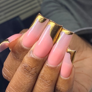 K&ouml;nnte beinhalten: Nahaufnahme einer Hand mit langen, quadratischen Acryln&auml;geln. Die N&auml;gel haben eine hellrosa Basis mit Goldfolienakzenten auf den Spitzen. Die Goldfolie hat ein strukturiertes, unregelm&auml;&szlig;iges Aussehen und verleiht Glamour.