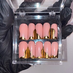 K&ouml;nnte beinhalten: Ein Set aus zehn k&uuml;nstlichen N&auml;geln in einer transparenten Kunststoffbox. Jeder Nagel ist zartrosa mit einem goldfarbenen, geschwungenen Design an der Basis. Die N&auml;gel sind rechteckig und scheinen zur Anwendung bereit zu sein.