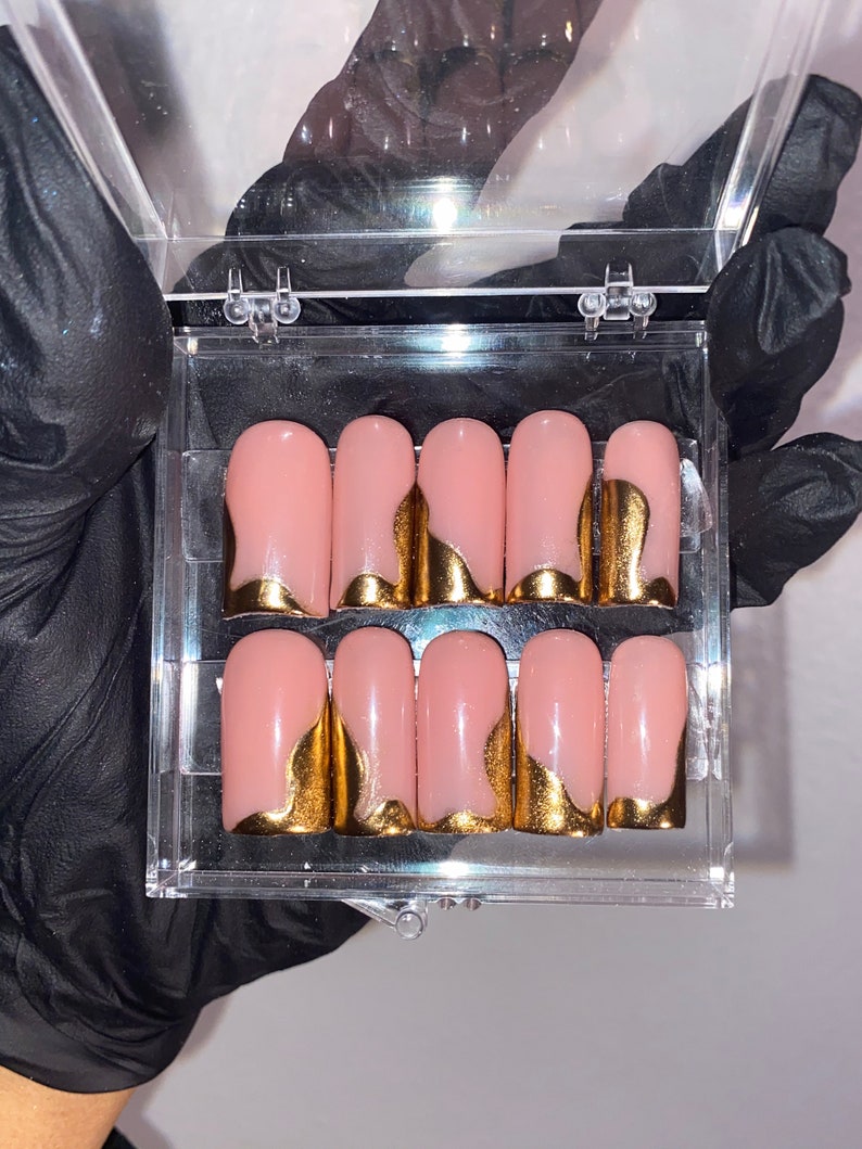 K&ouml;nnte beinhalten: Ein Set aus zehn k&uuml;nstlichen N&auml;geln in einer transparenten Kunststoffbox. Jeder Nagel ist zartrosa mit einem goldfarbenen, geschwungenen Design an der Basis. Die N&auml;gel sind rechteckig und scheinen zur Anwendung bereit zu sein.