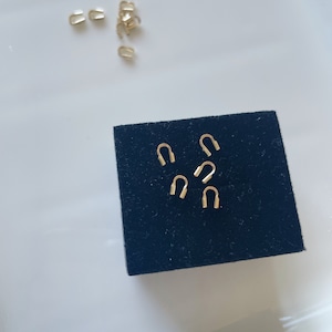 Peut inclure: Petites attaches de bijoux dorées, en forme d'agrafe ou de U, sont dispersées sur une surface blanche et un carré de velours noir. Ces attaches sont probablement utilisées en bijouterie pour relier des composants.