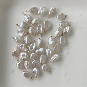 May include: A collection of white baroque pearls with gold jump rings. The pearls are various sizes and shapes, and they are arranged in a random pattern.