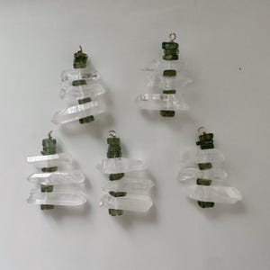May include: Five clear quartz crystal pendants with green stone accents and gold-coloured wire loops. The crystals are stacked in a tree-like design, with the green stones acting as the tree's foliage. The pendants are arranged on a light grey background.
