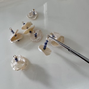 May include: Close-up of several handmade earrings. Each earring features a cluster of small, irregular, off-white petal-shaped elements, accented with a string of small, dark blue beads. The earrings have gold-coloured metal findings.