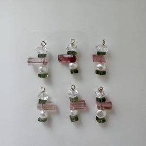 May include: Six handmade gemstone earrings with gold-colored hoops. Each earring features a combination of clear, pink, green, and white stones. The earrings are arranged on a white surface.