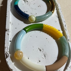May include: Two colorful bracelets with segmented designs rest on a white, textured tray. One bracelet features blue, yellow, and green sections, while the other combines light blue, yellow, and brown hues. Each bracelet has a central, spherical bead.