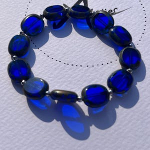 Könnte beinhalten: Ein Armband mit ovalen, tiefblauen Glasperlen. Die Perlen sind mit kleinen silberfarbenen Abstandshaltern verbunden. Das Armband wird auf einer weißen Oberfläche präsentiert, mit dem Text "mein Blumengarten" im Hintergrund.