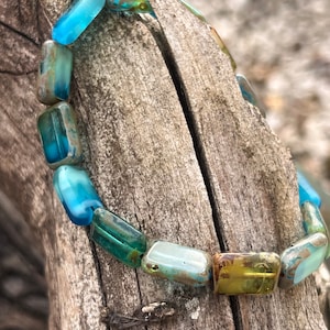 Könnte beinhalten: Ein Armband aus blauen, grünen und gelben Glasperlen. Die Perlen sind rechteckig und haben eine marmorierte, irisierende Oberfläche.